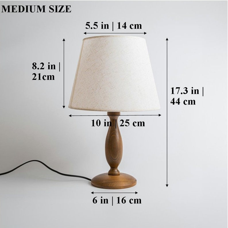 May include: A wooden table lamp with a beige fabric lampshade. The lamp is 17.3 inches tall, 10 inches wide, and has a 6-inch base. The lampshade is 5.5 inches wide and 8.2 inches tall.