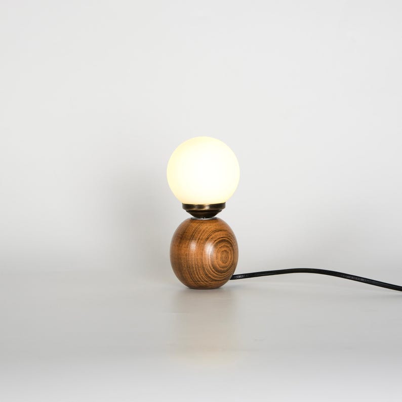May include: A contemporary table lamp featuring a spherical, warm white light bulb above a wooden base. The base consists of two wooden spheres. A black power cord extends from the base.