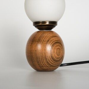 May include: A contemporary table lamp featuring a wooden sphere base and a frosted glass globe. The base is made of wood with visible grain patterns. The lamp has a brass-coloured metal connector and a black power cord.