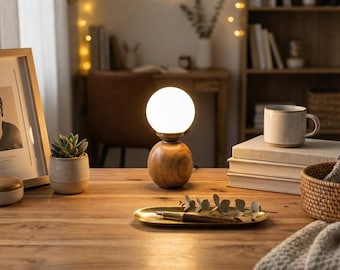 Puede incluir: Una lámpara de mesa de madera con una luz esférica blanca. La lámpara está sobre una base de madera con un veteado natural. La lámpara está sobre una mesa de madera con una foto enmarcada, una pequeña planta en maceta y una taza.