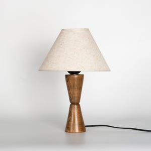 May include: A wooden table lamp with a beige fabric shade. The lamp base is made of two stacked, cone-shaped pieces of wood. A black power cord extends from the base. The lamp is set against a white background.