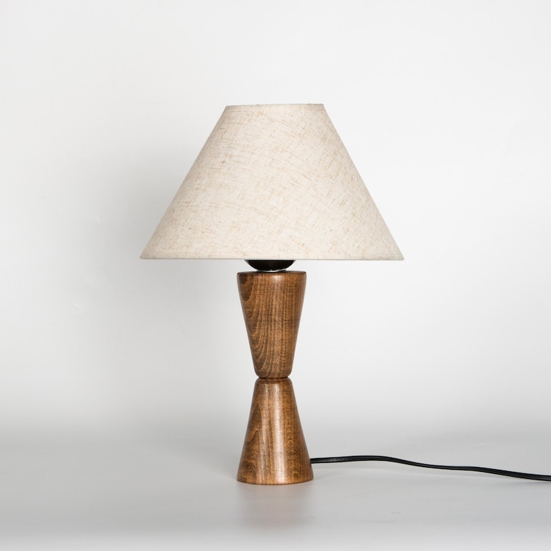 May include: A wooden table lamp with a beige fabric shade. The lamp base is made of two stacked, cone-shaped pieces of wood. A black power cord extends from the base. The lamp is set against a white background.