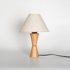May include: A wooden table lamp with a beige linen shade. The lamp base is made of light-colored wood and has a unique hourglass shape. A black power cord extends from the base. The lamp is on a white surface.