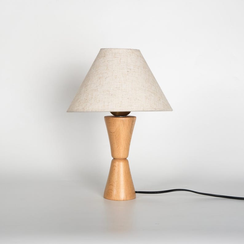 May include: A wooden table lamp with a beige linen shade. The lamp base is made of light-colored wood and has a unique hourglass shape. A black power cord extends from the base. The lamp is on a white surface.