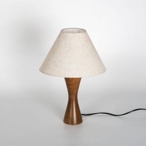 May include: A small wooden table lamp with a beige fabric lampshade. The lamp has a hourglass-shaped wooden base and a conical lampshade. A black power cord extends from the base.
