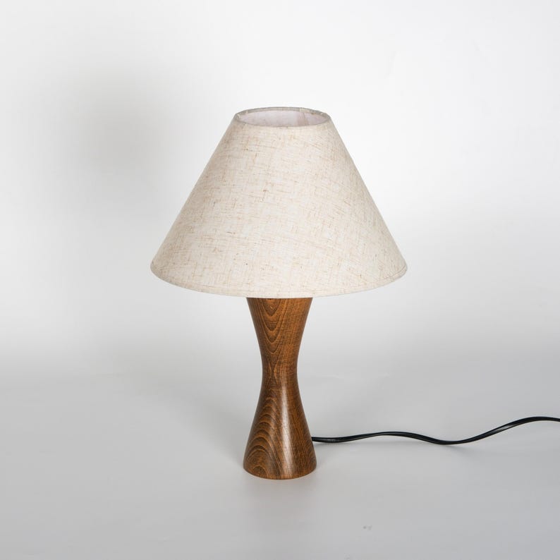 May include: A small wooden table lamp with a beige fabric lampshade. The lamp has a hourglass-shaped wooden base and a conical lampshade. A black power cord extends from the base.