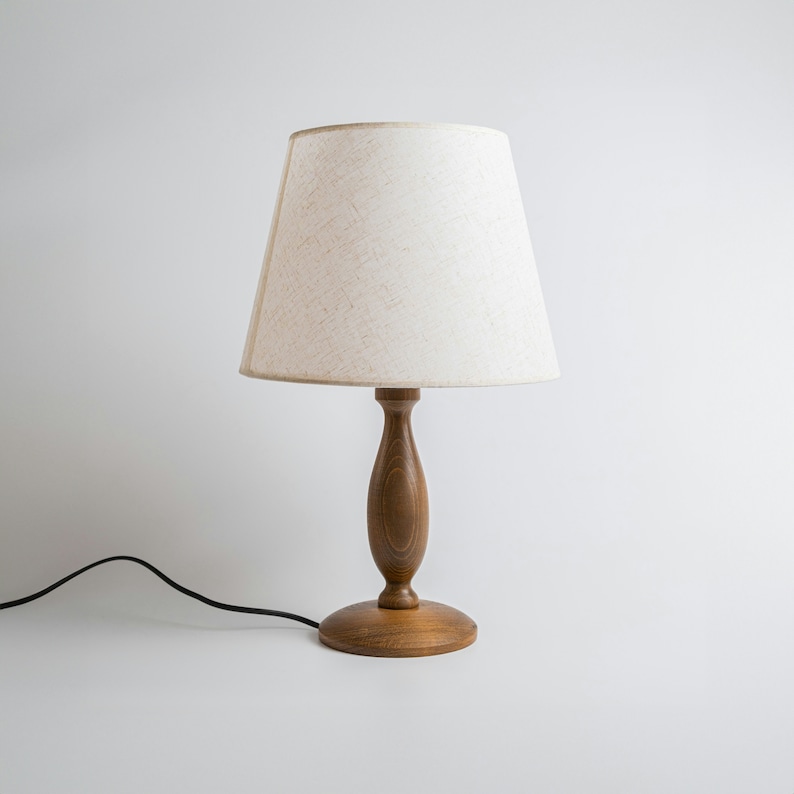 May include: A wooden table lamp with a white linen lampshade. The lamp has a simple, elegant design and would look great in any home.