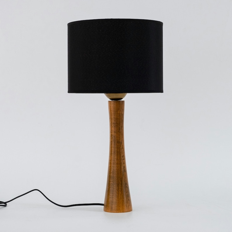 May include: A wooden table lamp with a black fabric lampshade. The lamp has a tall, slender base with a natural wood finish.
