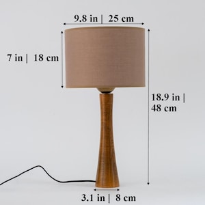 May include: A wooden table lamp with a brown fabric lampshade. The lamp is 18.9 inches tall and the lampshade is 9.8 inches wide. The base of the lamp is 3.1 inches wide.