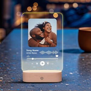 May include: A rectangular clear acrylic plaque with a light-up edge, featuring a printed photograph of a smiling couple. The display includes a music player interface with text reading Song Name and Artist Name, a waveform, and playback controls.