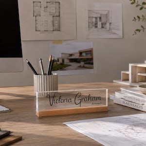 May include: A desk nameplate with the name "Victoria Graham" and the title "Architect" in black script. The nameplate is clear acrylic with a wooden base. The desk also has a computer, pens, and architectural drawings.