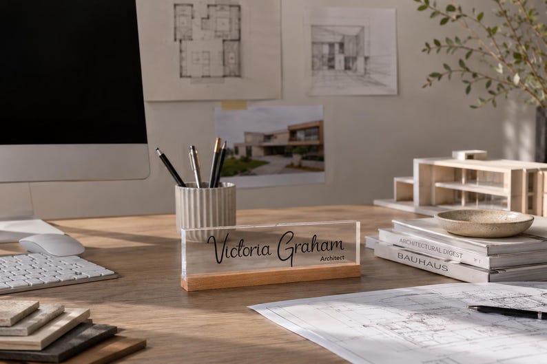 May include: A desk nameplate with the name "Victoria Graham" and the title "Architect" in black script. The nameplate is clear acrylic with a wooden base. The desk also has a computer, pens, and architectural drawings.