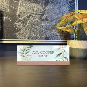 May include: A desk nameplate with the text "AVA COOPER Dentist" in black font. The nameplate has a light blue background with green and gold leaf accents. It sits on a wooden base. A map and a plant are in the background.
