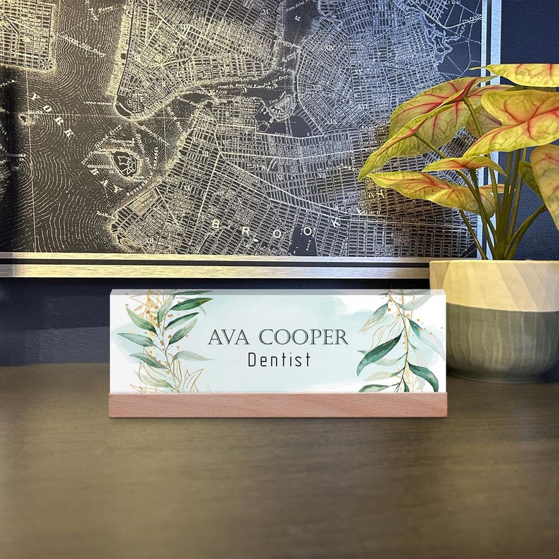 May include: A desk nameplate with the text "AVA COOPER Dentist" in black font. The nameplate has a light blue background with green and gold leaf accents. It sits on a wooden base. A map and a plant are in the background.