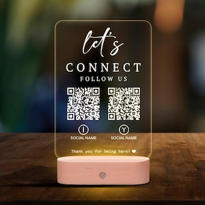 May include: A clear rectangular acrylic sign with rounded corners, featuring the text "let's CONNECT FOLLOW US", two QR codes, two circular icons with letters "I" and "Y", "SOCIAL NAME" labels, and "Thank you for being here!" on a light wood base.