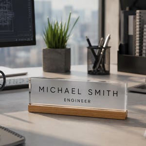 May include: A clear acrylic desk nameplate with the name "MICHAEL SMITH" and the title "ENGINEER" in black lettering. The nameplate sits on a light-colored wooden base. A mug, pens, and a laptop are also on the desk.