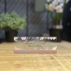 May include: A clear acrylic desk nameplate with a floral design and gold accents. The name "Abigail Cook" and the title "Senior Manager" are printed in black on the nameplate. The nameplate has a wooden base.