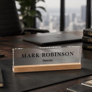 May include: A clear acrylic desk nameplate with the name "MARK ROBINSON" and the title "Director" in black text. The nameplate sits on a light wood base. The desk is dark wood and has a black leather desk pad.