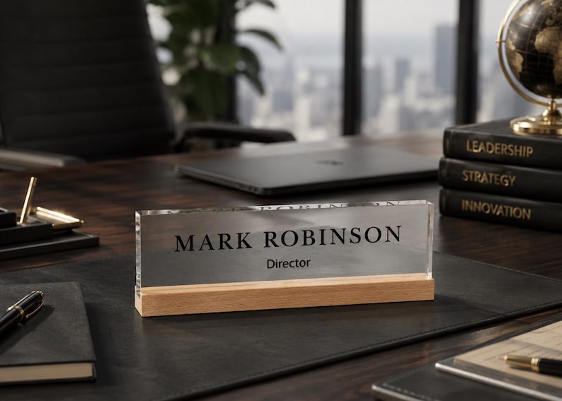 May include: A clear acrylic desk nameplate with the name "MARK ROBINSON" and the title "Director" in black text. The nameplate sits on a light wood base. The desk is dark wood and has a black leather desk pad.