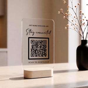 May include: A clear rectangular acrylic sign with rounded corners, featuring the black text GET MORE WITH OUR APP, Stay connected, and SCAN TO DOWNLOAD, along with a central QR code. The sign is mounted on a light-colored wooden base.