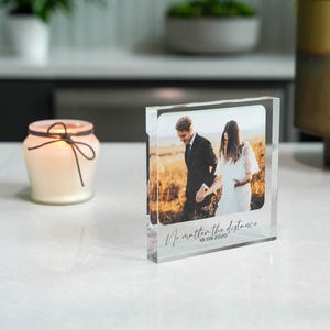 May include: A clear acrylic photo block featuring a couple holding hands, with the text "No matter the distance 18.09.2020". The photo block is on a white surface, with a candle in a glass jar in the background.
