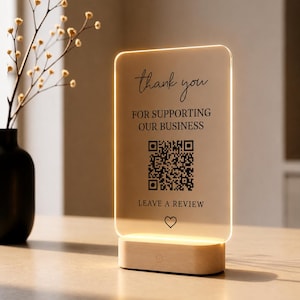 May include: A rectangular clear acrylic sign with rounded corners, illuminated by a light-colored wooden base. The sign features black text that reads thank you FOR SUPPORTING OUR BUSINESS, a QR code, LEAVE A REVIEW, and a small heart icon.
