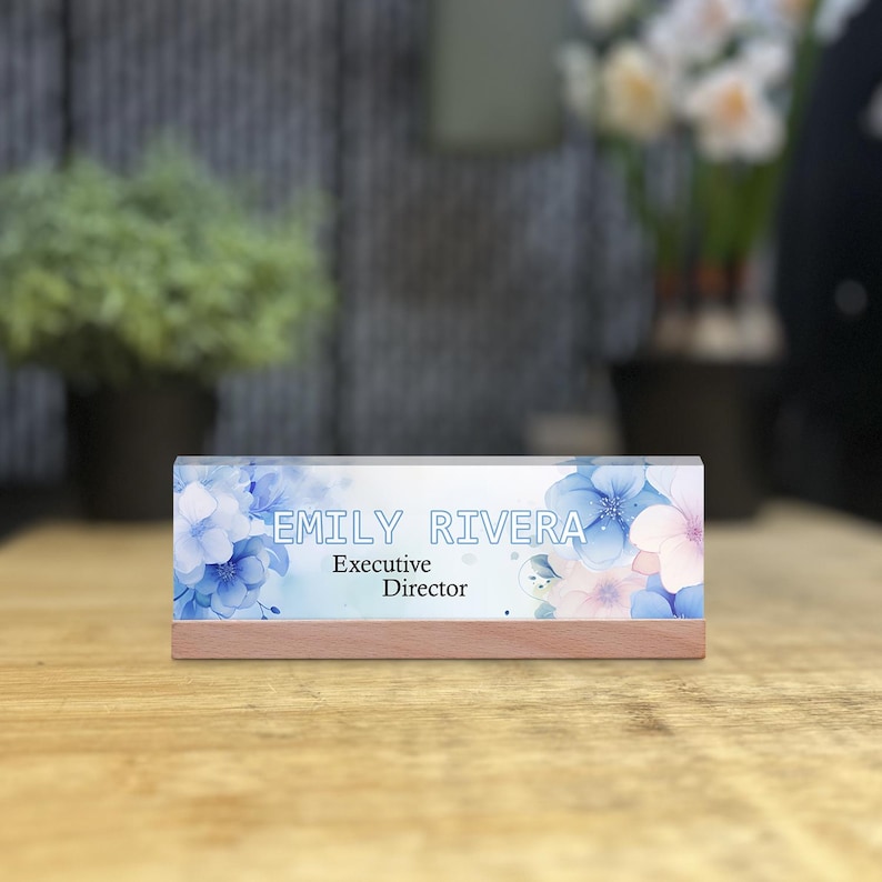 May include: A personalized desk nameplate with the name "EMILY RIVERA" and title "Executive Director" in white text. The nameplate features a floral design with blue and pink flowers, set on a wooden base.