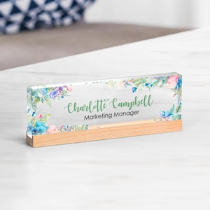 May include: A clear acrylic desk nameplate with floral accents and a wooden base. The nameplate features the name "Charlotte Campbell" and the title "Marketing Manager" in green script.
