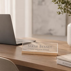 May include: A desk nameplate with the text "Sarah Bennet, Chief Executive Officer" sits on a wooden desk. A laptop, a vase with greenery, a book, and a pen are also on the desk. The nameplate is clear acrylic with a wooden base.