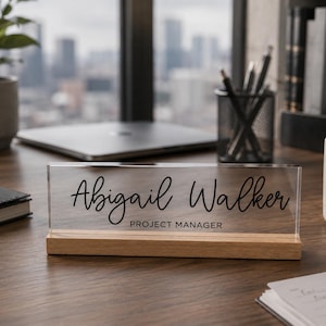 May include: A clear acrylic desk nameplate with the name "Abigail Walker" and the title "Project Manager" in black script. The nameplate sits on a wooden base. Other items on the desk include a laptop, a mug, and a potted plant.