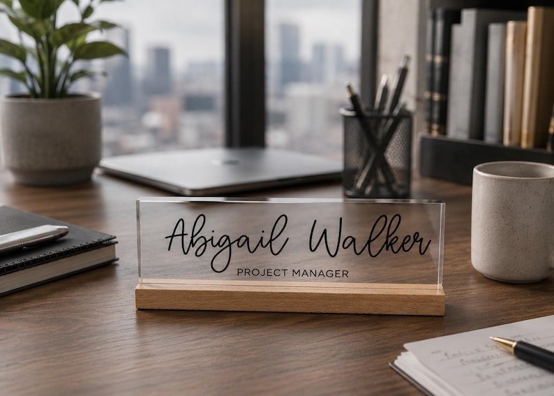 May include: A clear acrylic desk nameplate with the name "Abigail Walker" and the title "Project Manager" in black script. The nameplate sits on a wooden base. Other items on the desk include a laptop, a mug, and a potted plant.