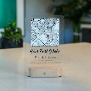 May include: A personalized acrylic map light with a wooden base. The map shows a street layout with a highlighted red location. Text reads "Our First Date", names, and a date. The base has a small, round light.
