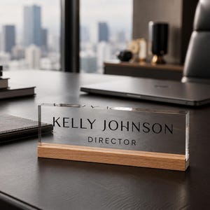 May include: A clear acrylic desk nameplate with the name "KELLY JOHNSON" and the title "DIRECTOR" in black lettering. The nameplate sits on a wooden base. The background includes a desk, a laptop, and a cityscape.