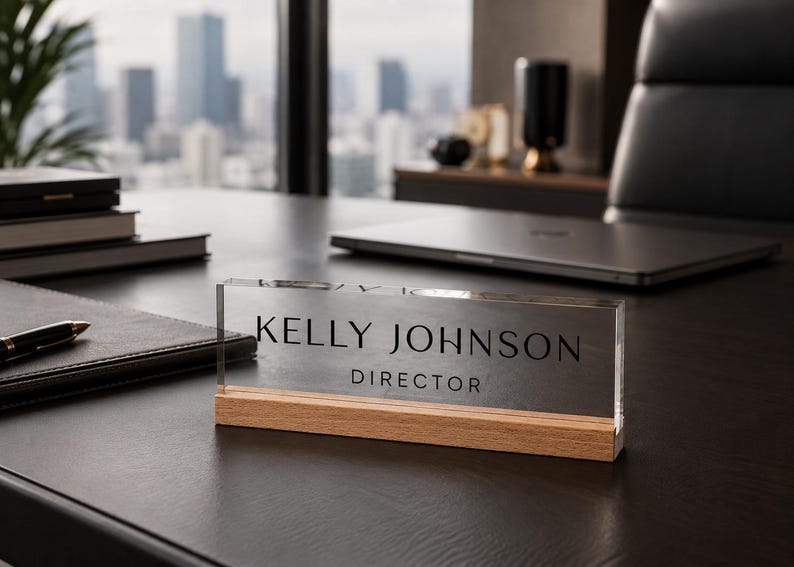 May include: A clear acrylic desk nameplate with the name "KELLY JOHNSON" and the title "DIRECTOR" in black lettering. The nameplate sits on a wooden base. The background includes a desk, a laptop, and a cityscape.