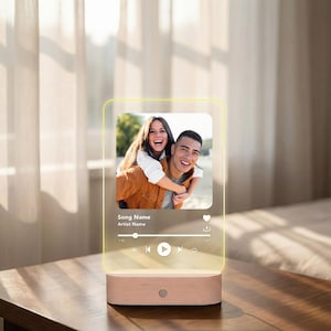 May include: A personalized night light featuring a photo of a couple, resembling a music player interface. The light has a wooden base and a clear acrylic panel with the photo and song details. The text on the panel reads "Song Name" and "Artist Name".