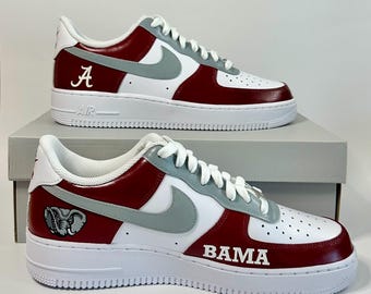 May include: A pair of white, crimson, and grey low-top trainers featuring a grey Nike swoosh, white laces, and white soles. The shoes display the letter A, an elephant graphic, and the text BAMA in white lettering on the crimson side panels.