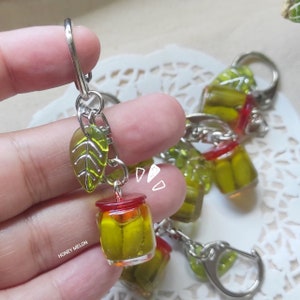 Puede incluir: Un conjunto de cinco llaveros de tarro de vidrio verde con tapas rojas. Cada llavero tiene una pequeña hoja verde y un anillo de llave plateado. Los llaveros están dispuestos sobre un fondo de encaje blanco.