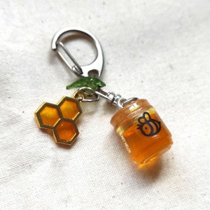 Può includere: Un piccolo barattolo di vetro pieno di miele con un design a forma di ape, attaccato a un portachiavi argentato con un ciondolo a forma di favo dorato. Il barattolo ha la scritta "Honey Melon".