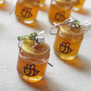 May include: A pair of honey jar earrings with a bee design. The jars are made of clear glass with a gold lid and filled with a honey-colored liquid. The earrings are attached to silver hooks.