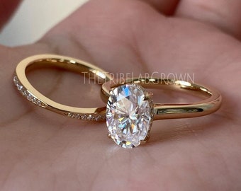 May include: A gold engagement ring set featuring an oval-cut diamond solitaire and a matching band with small accent stones. The rings are displayed on a neutral background, showcasing their polished finish and intricate design.