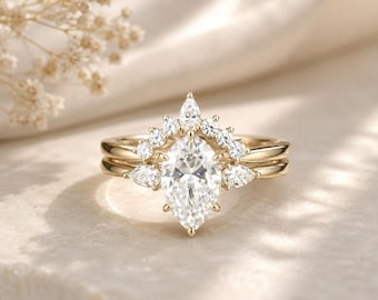 May include: A gold ring set featuring a central marquise-cut clear stone flanked by two smaller pear-cut stones. A curved band sits above the main stone, adorned with a series of round-cut clear stones arranged in a crown-like formation.
