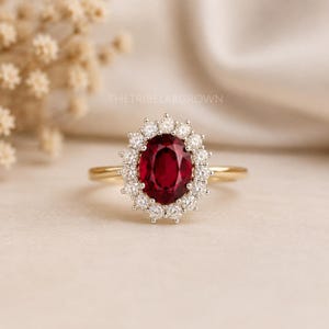 Puede incluir: Un anillo de oro con una gran piedra preciosa de color rojo intenso y corte ovalado en el centro, rodeada por un halo de pequeñas piedras blancas claras y redondas. La banda es fina y de oro pulido. El texto THETRIBELABGROWN es visible arriba.