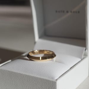 Op de afbeelding: Een goudkleurige ring met een facetgeslepen ontwerp, in een open witte sieradendoos. De ring heeft een geometrische, hoekige vorm. De merknaam is zichtbaar op de doos.