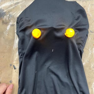 Puede incluir: Una tela negra drapeada sobre una forma invisible, con dos globos amarillos brillantes y luminosos colocados para parecer ojos. Los globos son redondos y emiten una luz naranja cálida, creando un efecto espeluznante.