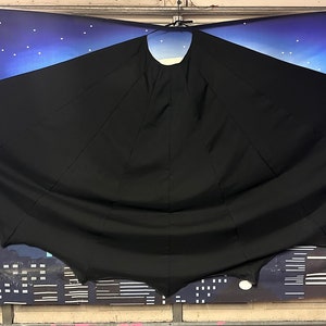 May include: A black cape with a large, rounded collar and a pointed hood. The cape is hanging against a backdrop of a city skyline and a starry night sky.