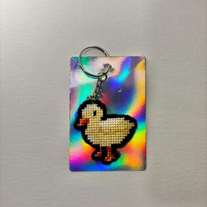 May include: A handmade keychain featuring a cross-stitched duckling design. The duckling is yellow with a black outline and orange beak and feet. The keychain is attached to a holographic card with a silver keyring.