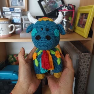 Puede incluir: Un toro azul de crochet con detalles florales coloridos y un borla rojo, naranja y azul. El toro sostiene una pequeña borla rojo, naranja y azul.