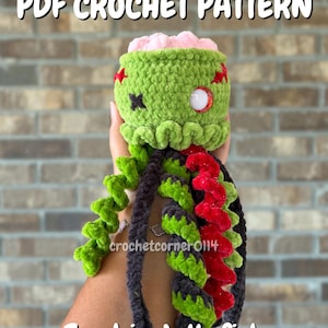 May include: A PDF crochet pattern for a zombie jellyfish. The plush features a green head with a pink brain top, one white eye, and a black stitched eye. It has long, spiraled tentacles in shades of green, black, and red. Text reads PDF CROCHET PATTERN, crochetcorner0114, and Zombie Jellyfish.