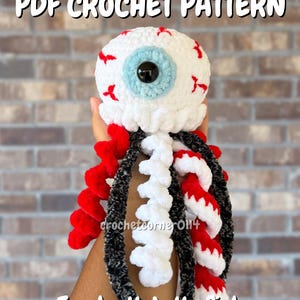 May include: A crochet jellyfish toy featuring a large white spherical head with red vein details, a light blue iris, and a black pupil. The jellyfish has multiple spiraling tentacles in solid red, solid white, and a textured black yarn.