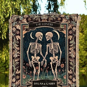 May include: A decorative throw blanket featuring a dark background with two skeletons holding hands. The words "THE LOVERS" and "DYLAN & GABBY 2024" are displayed on the blanket. The blanket has a floral border and fringe.